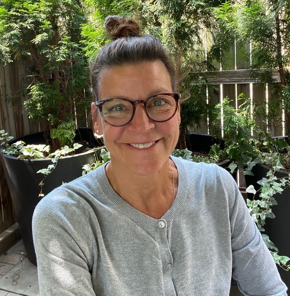 A smiling woman wearing glasses and a grey cardigan, with her hair done up in a bun. The photo is taken in a sunny yard, against a fence covered in foliage.
