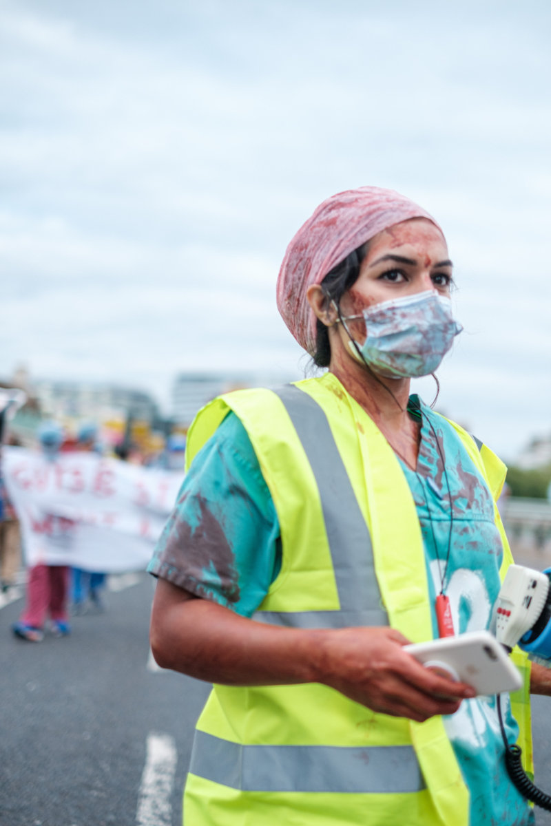A woman nurse wears a blue gown stained with red during a street protest.