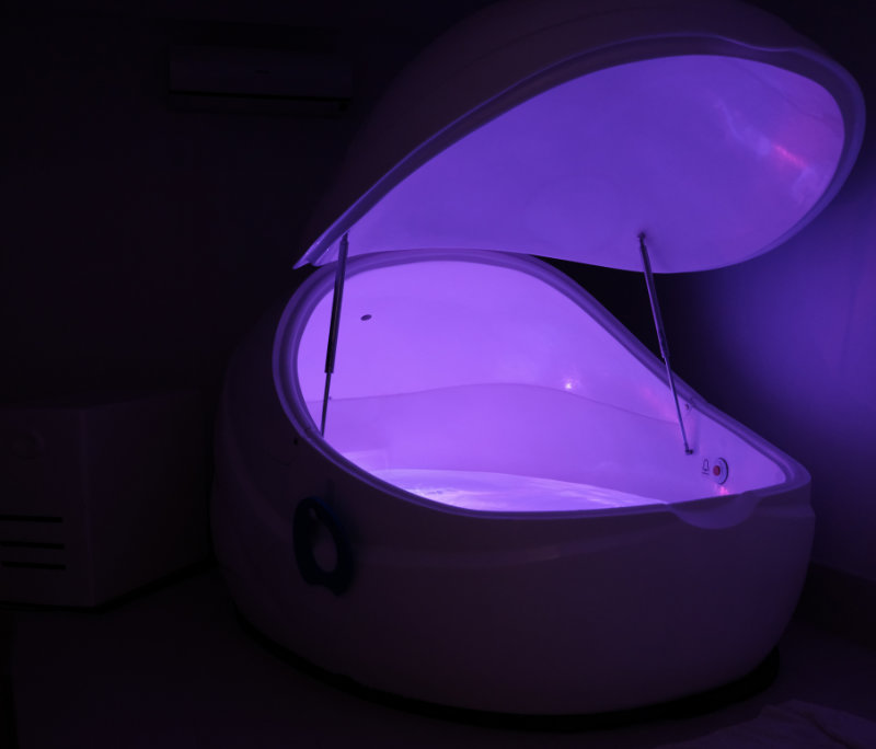 In the dark sits a purple-lit sensory deprivation tank with a top lid open above it showing the water within.