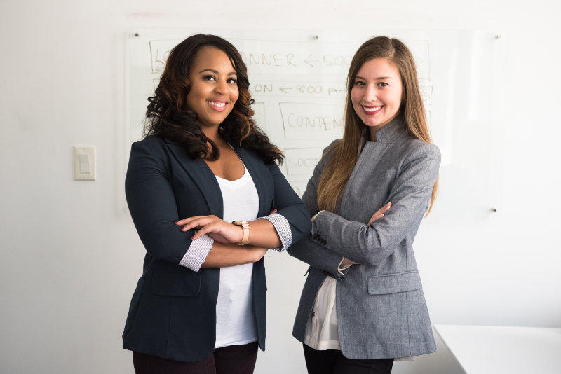 Two women posing next to each other in an office. The black woman is wearing a dark suit and the white woman is wearing a grey suit.